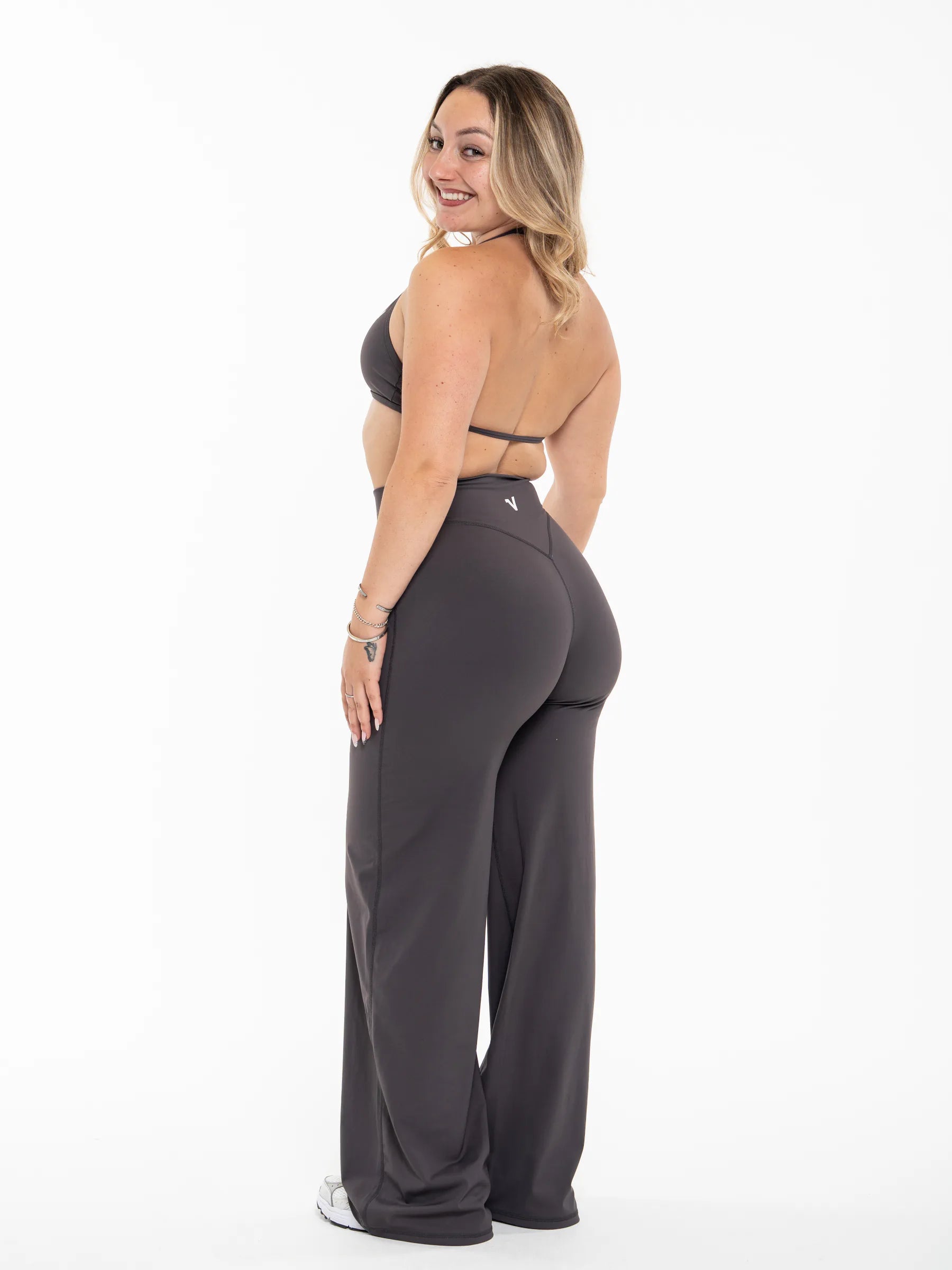 Veritone Sculpt Flared Pants, Charcoal Grey – A woman modeling dark gray wide-leg athletic pants with a matching sports bra, shown from a back-three-quarter angle against a plain white studio background.