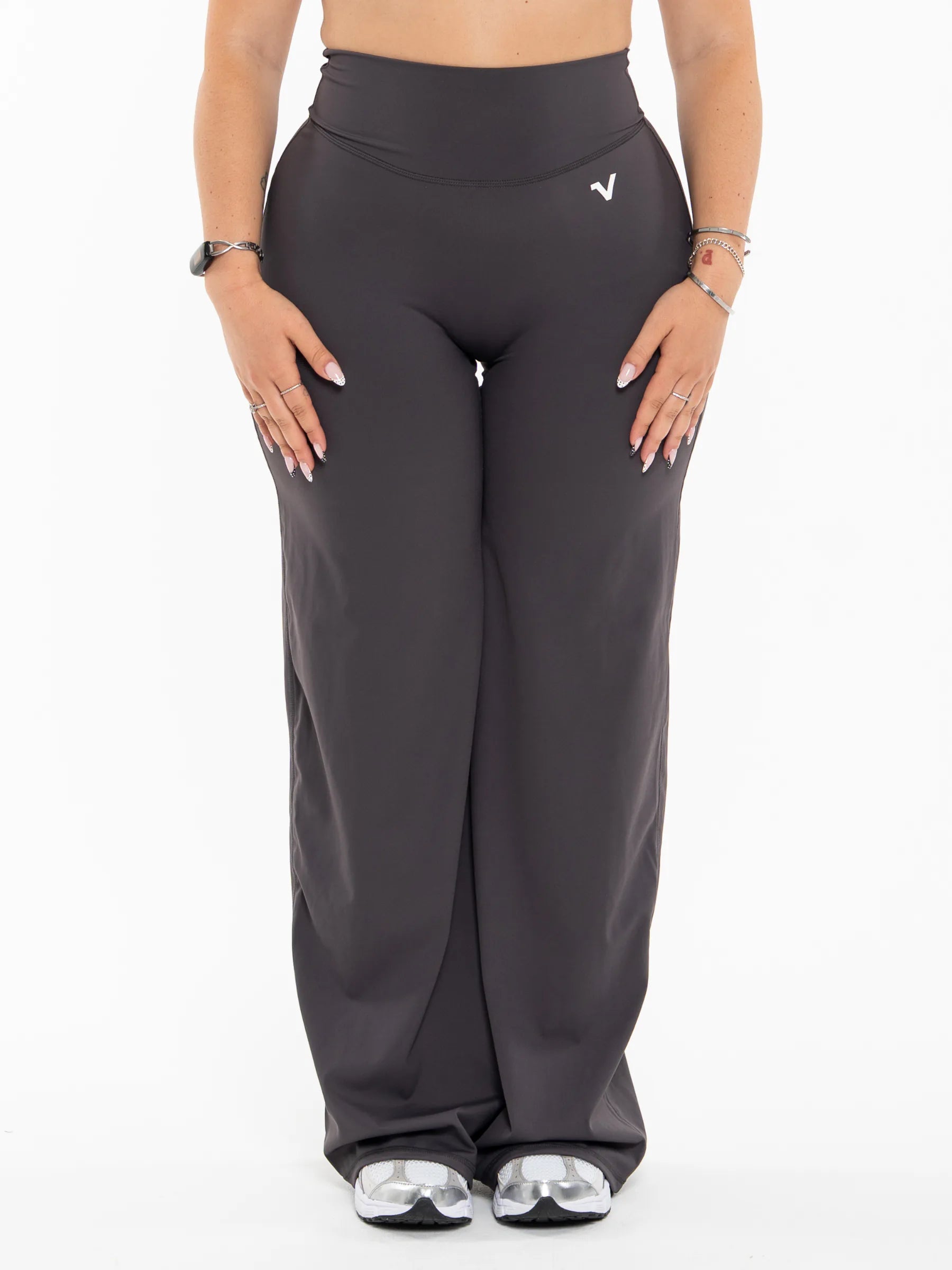 Veritone Sculpt Flared Pants, Charcoal Grey – A model wearing dark gray wide-leg athletic pants with a small white stylized logo on the left hip, shown from the waist down on a white background.