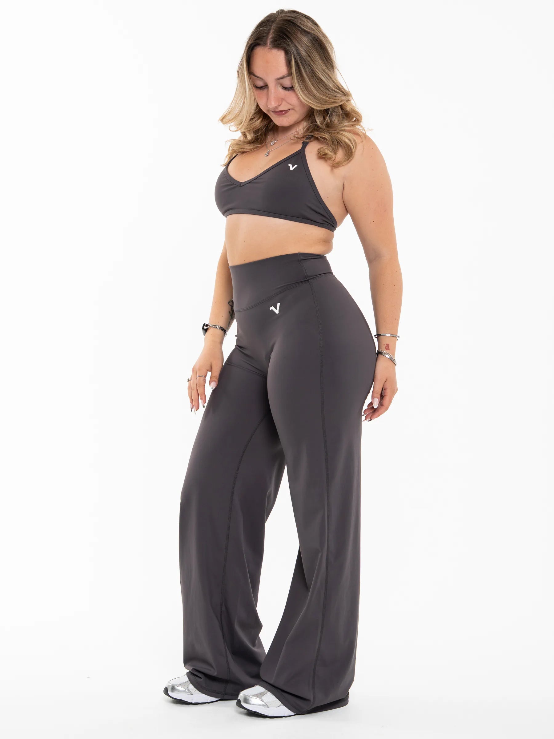 Veritone Sculpt Flared Pants, Charcoal Grey – A woman modeling a dark gray athletic set consisting of a sports bra and high-waisted wide-leg pants, shown against a plain white studio background.