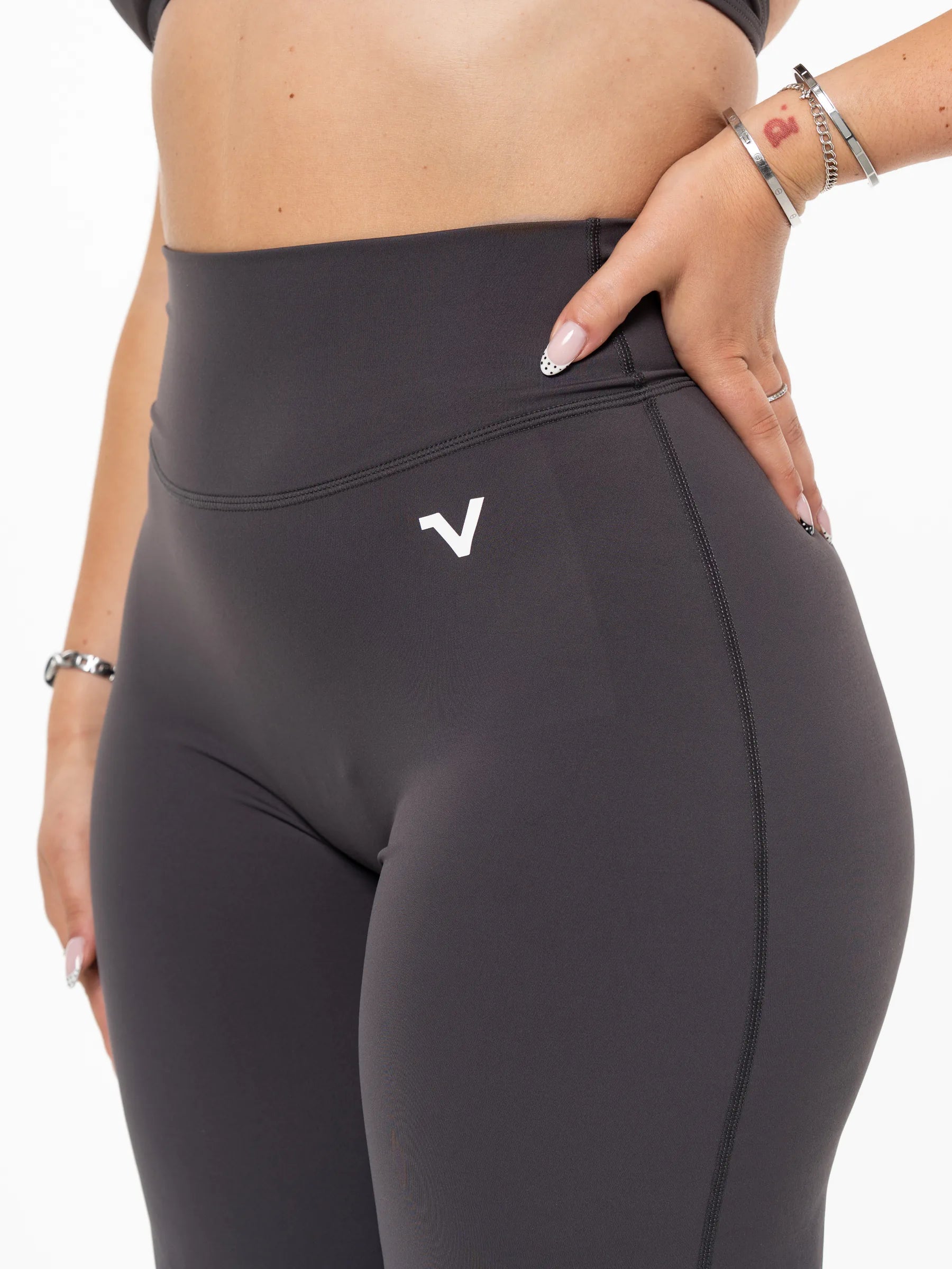 Veritone Sculpt Flared Pants, Charcoal Grey – Close-up of a model wearing dark gray high-waisted athletic leggings with a white stylized 'V' logo on the hip, set against a plain white background.