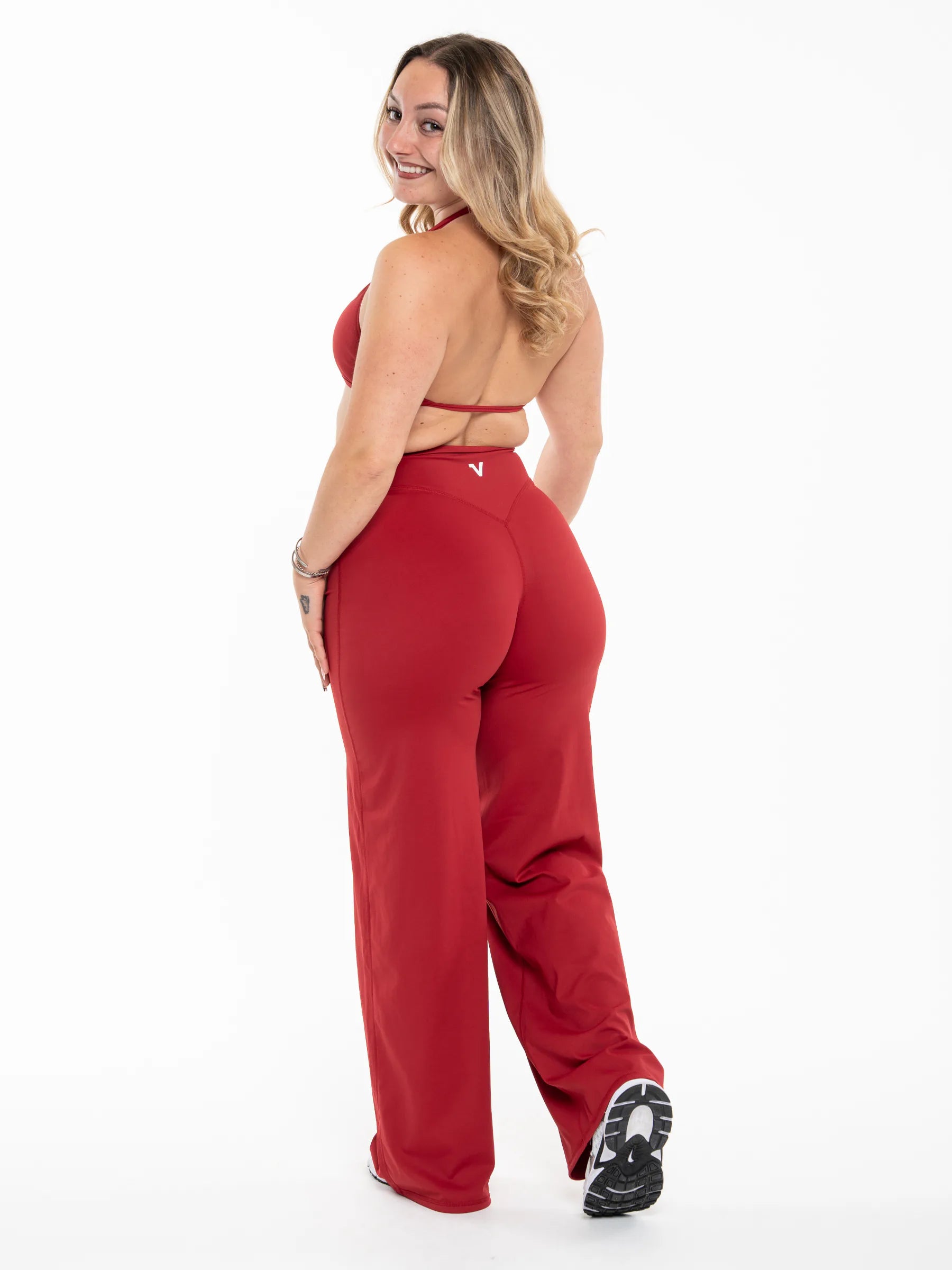 Veritone Sculpt Flared Pants, Crimson Red – A woman modeling a matching red two-piece activewear set, featuring a halter-style sports bra and high-waisted, wide-leg pants, shown from a rear three-quarter angle against a white studio background.