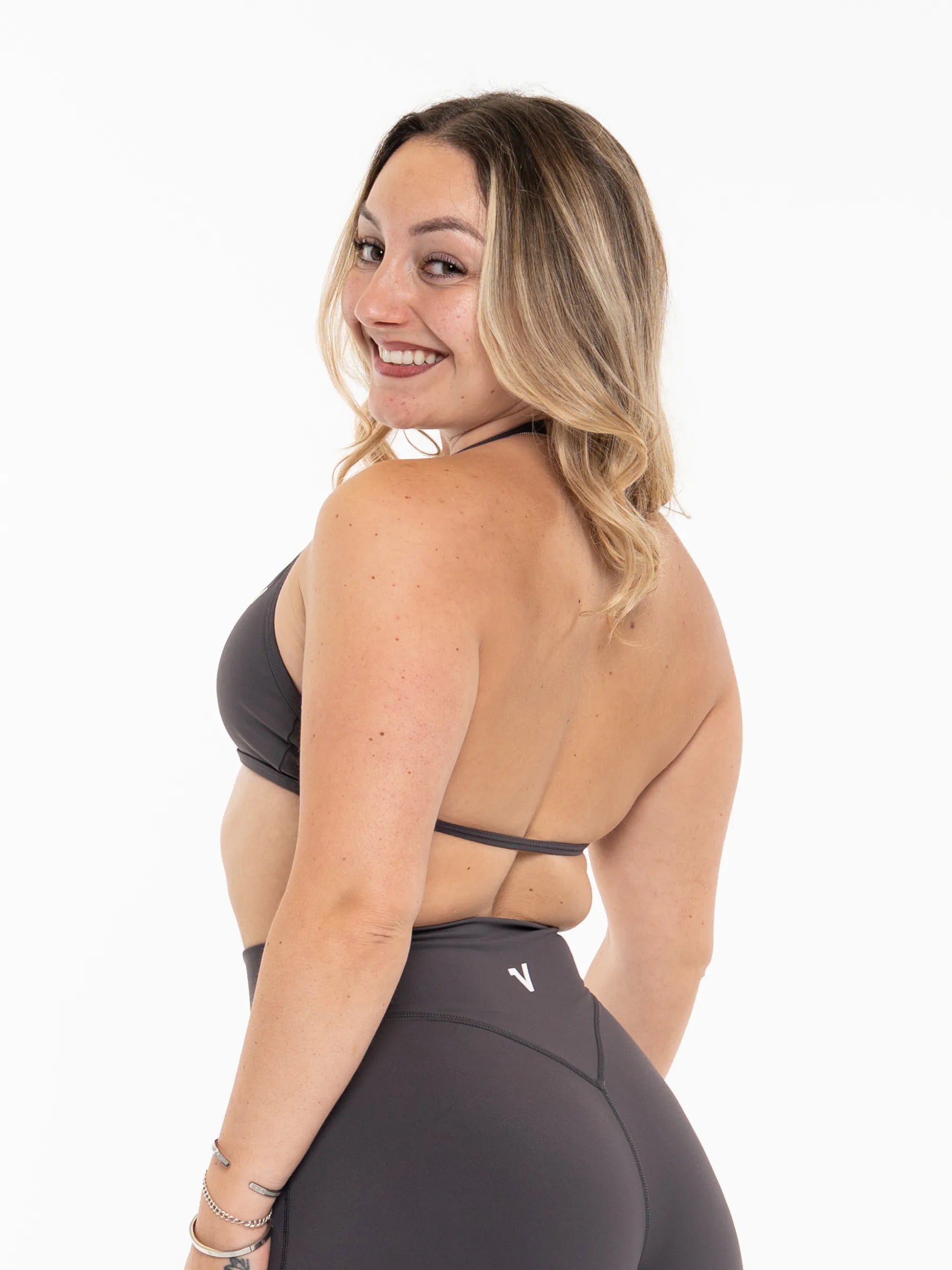 Veritone Sculpt Sports Bra, Charcoal Grey – A woman with blonde-highlighted hair is shown from the back wearing a dark gray sports bra and matching high-waisted leggings with a small white logo, set against a plain white background.