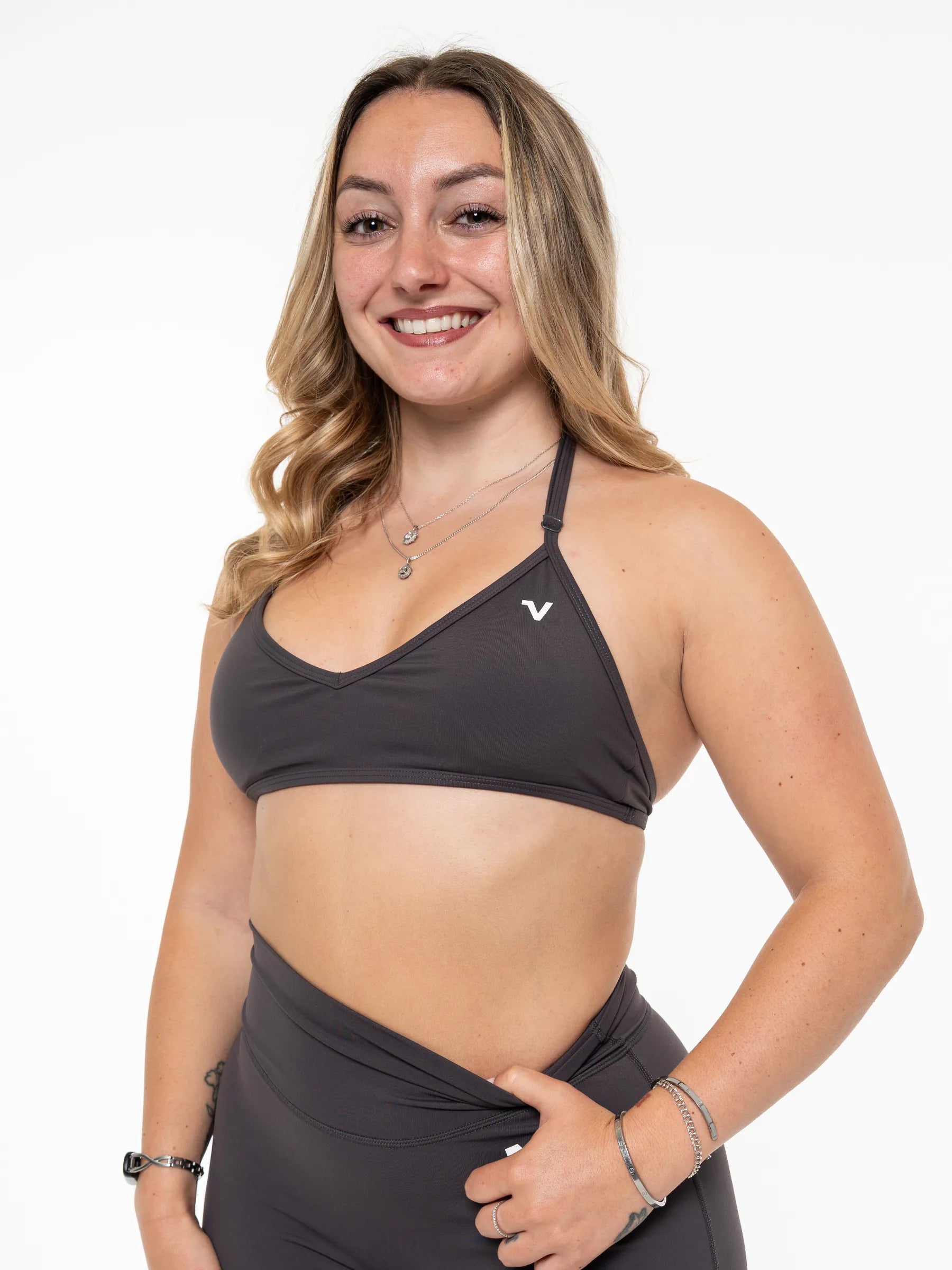 Veritone Sculpt Sports Bra, Charcoal Grey – A smiling woman with blonde hair wearing a dark gray sports bra and matching high-waisted leggings, posing against a solid white studio background.
