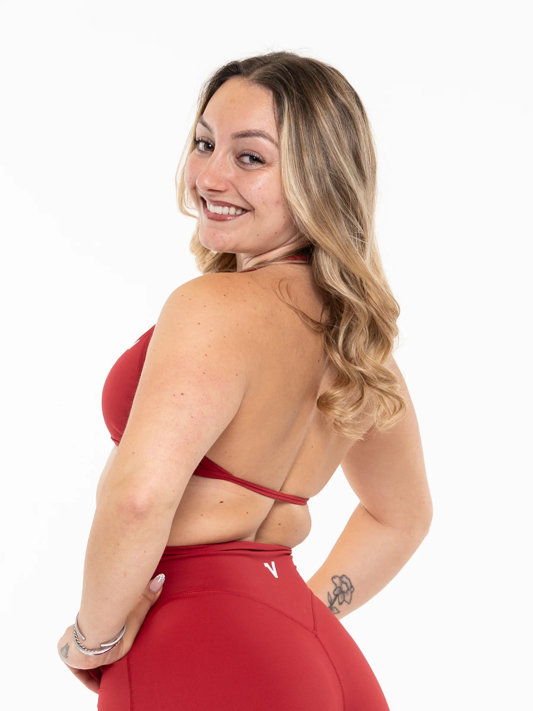 Veritone Sculpt Sports Bra, Crimson Red – A woman with blonde wavy hair is shown from the back, wearing a red strappy sports bra and matching red high-waisted leggings against a plain white background.