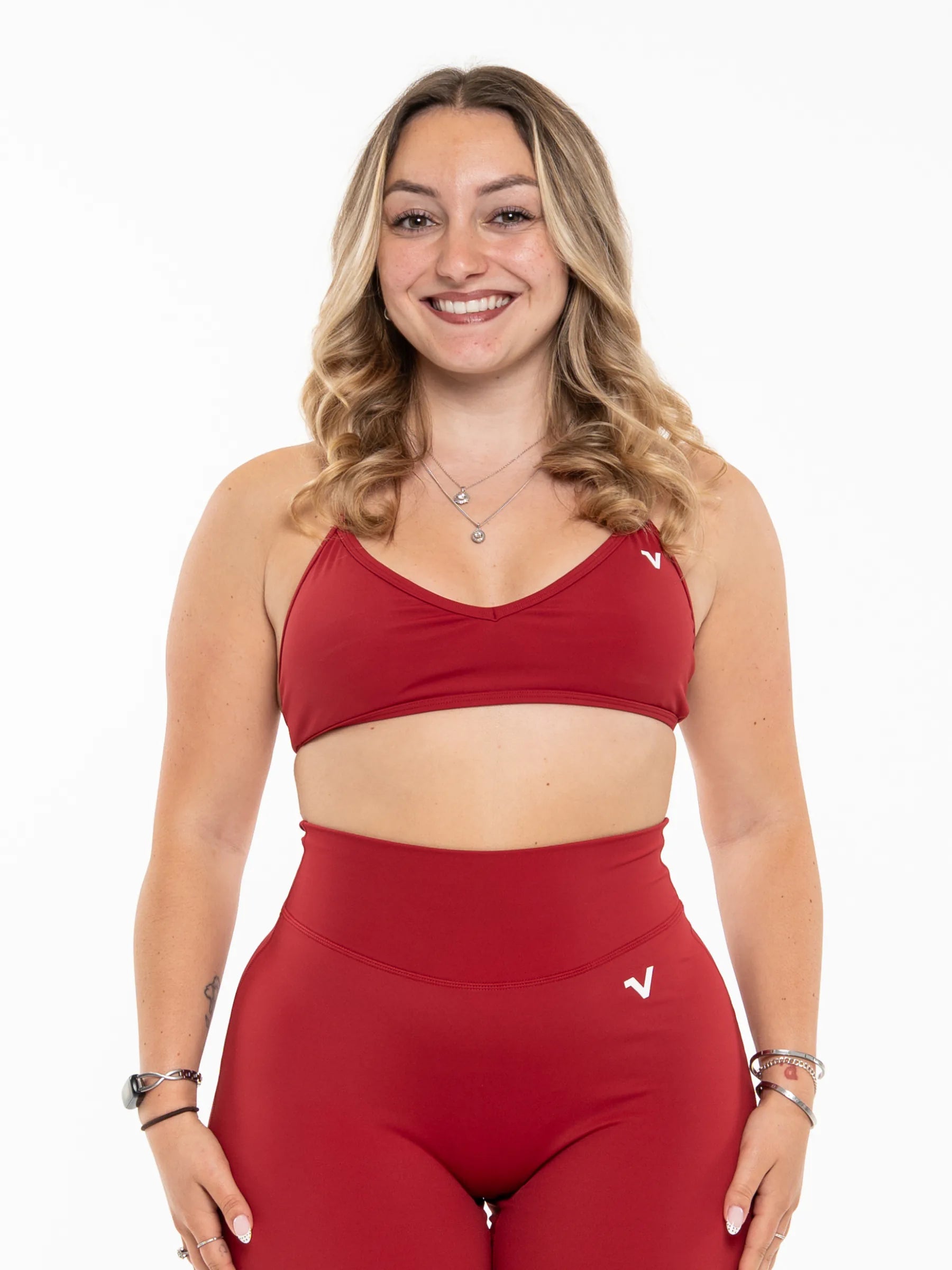 Veritone Sculpt Sports Bra, Crimson Red – A smiling woman with blonde wavy hair models a matching red activewear set consisting of a sports bra and high-waisted leggings, set against a plain white background.